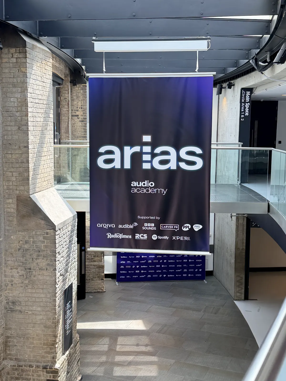 An ARIAS banner listing the sponsors hanging above the stairs in the Roundhouse foyer, between old brick walls and a glass balcony.