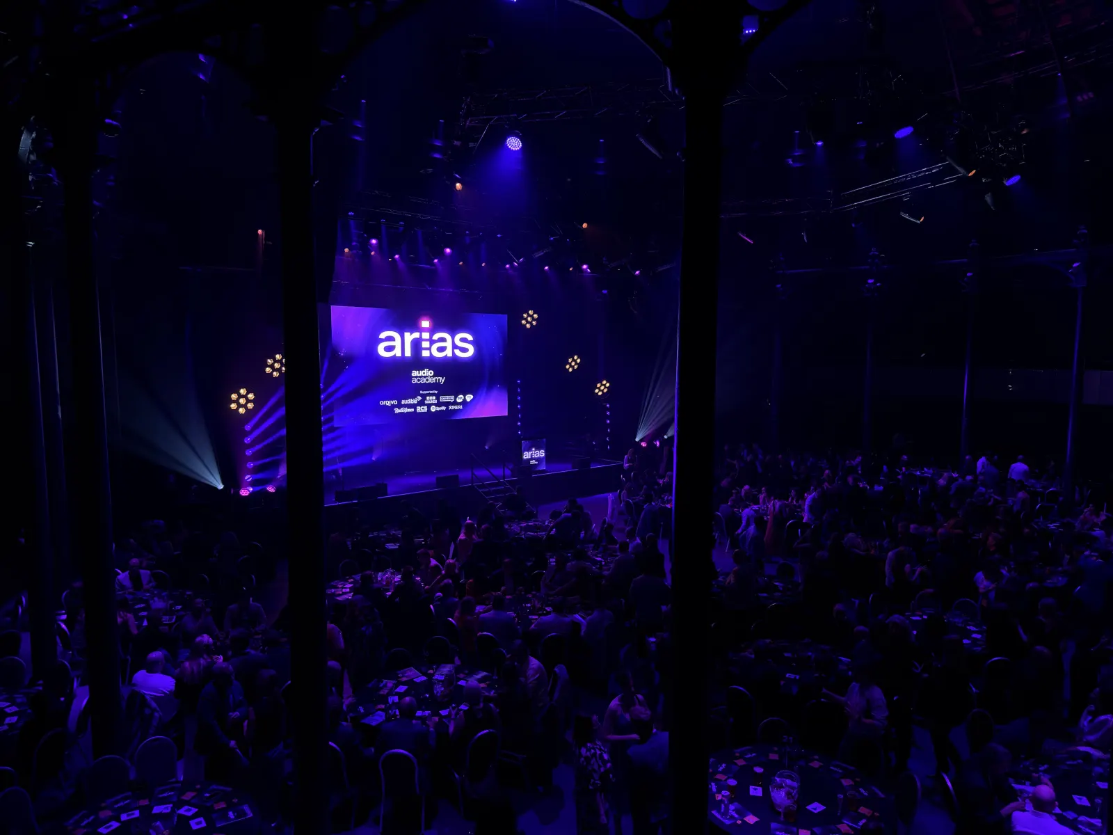 The ARIAS ceremony seen between the Roundhouse's iron columns, a full room of tables in the dark facing a stage lit blue and purple.