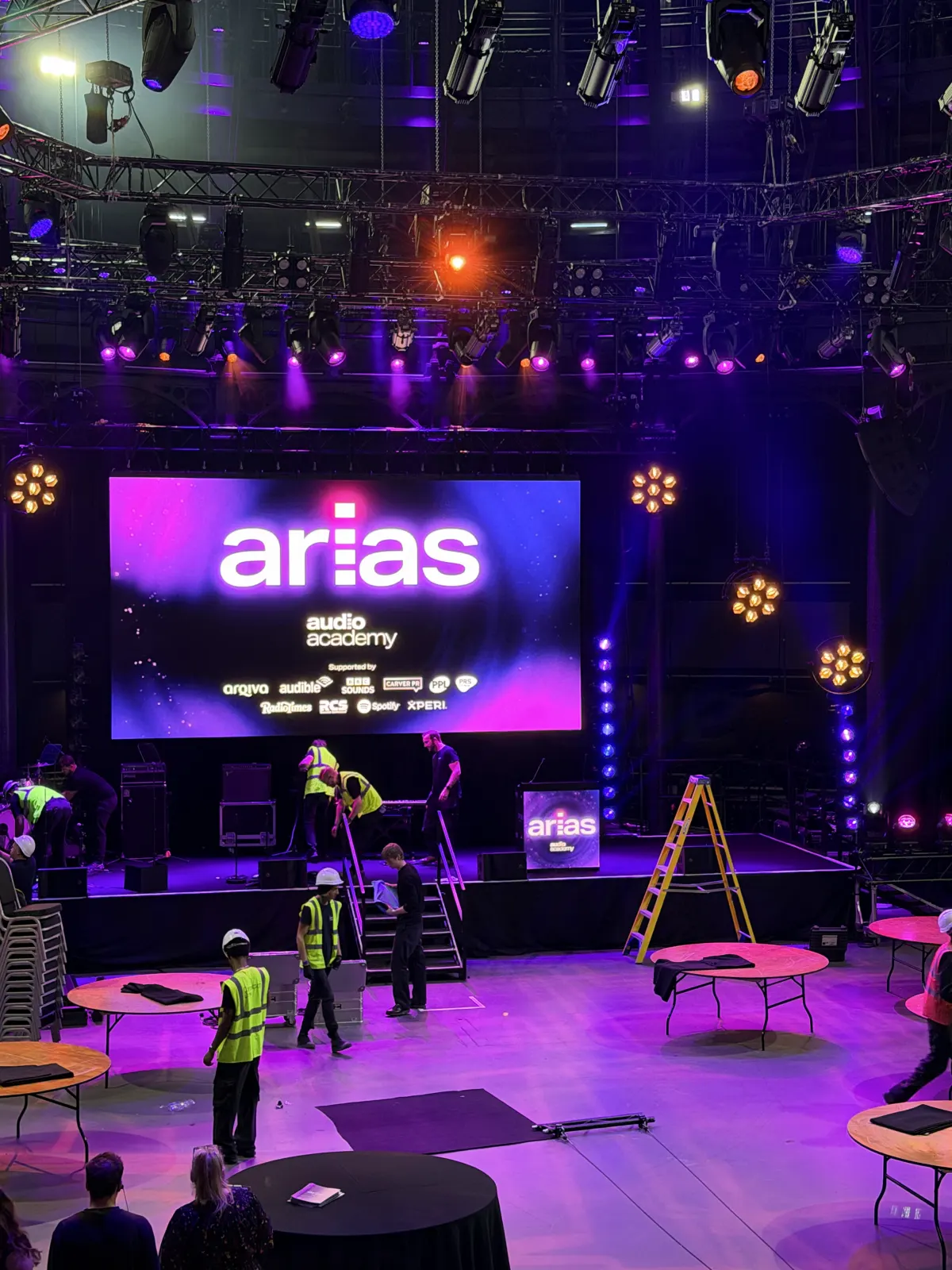 The Roundhouse main space in purple light during the afternoon setup, crew in hi-vis carrying equipment between bare round tables in front of the stage.