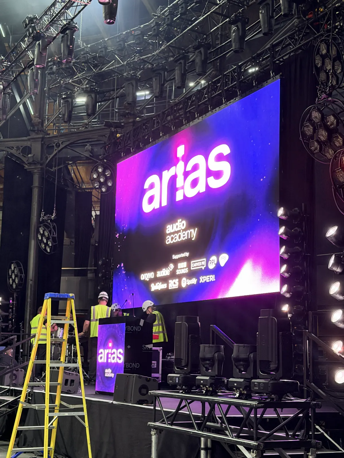 Crew in hard hats and hi-vis building the ARIAS stage under a lighting rig, a yellow stepladder in front of a big purple screen showing the arias and Audio Academy logos.