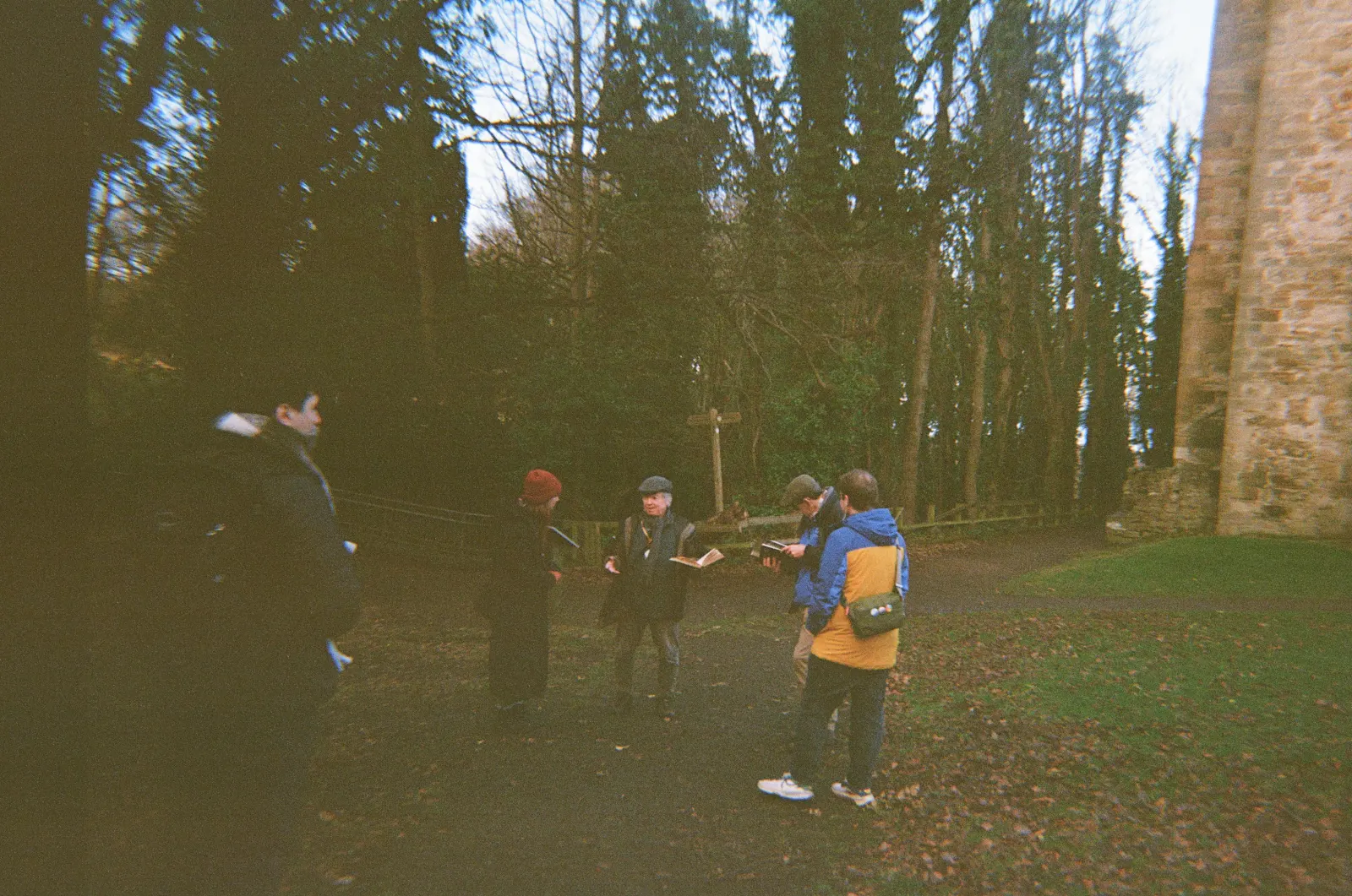 Adam Stoner, Sarah MacGillivray, Mark Watson from Historic Scotland and Ian Sheerer from Kinneil House gathered on a path among trees at Kinneil House, one of them in a flat cap reading from an open book, the sandstone wall of the house at the right edge.