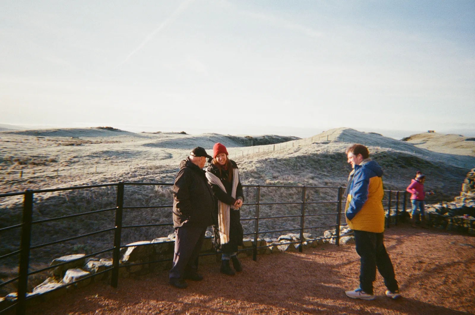 Sarah MacGillivray, Julia Hasse, Adam Stoner and an interviewee at black railings on a red gravel path, rolling hills behind them white with frost.