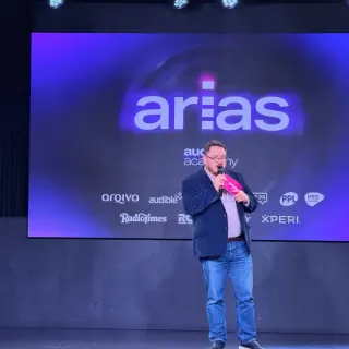 A man on stage at the ARIAS, speaking into a pink microphone in front of a large screen showing the arias and Audio Academy logos.
