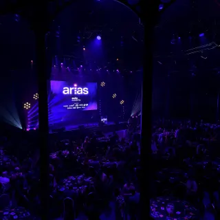 The ARIAS ceremony seen between the Roundhouse's iron columns, a full room of tables in the dark facing a stage lit blue and purple.