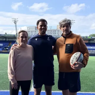 Three people arm in arm on the pitch at Cardiff Arms Park, one in Cardiff Rugby kit holding a Gilbert rugby ball, under a bright blue sky.