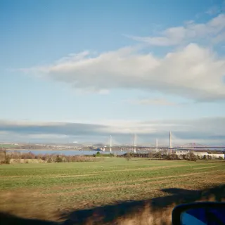 Taken from a moving car, green fields running down to the Firth of Forth, the white towers of the Queensferry Crossing and the red Forth Bridge on the horizon, a wing mirror in the corner.